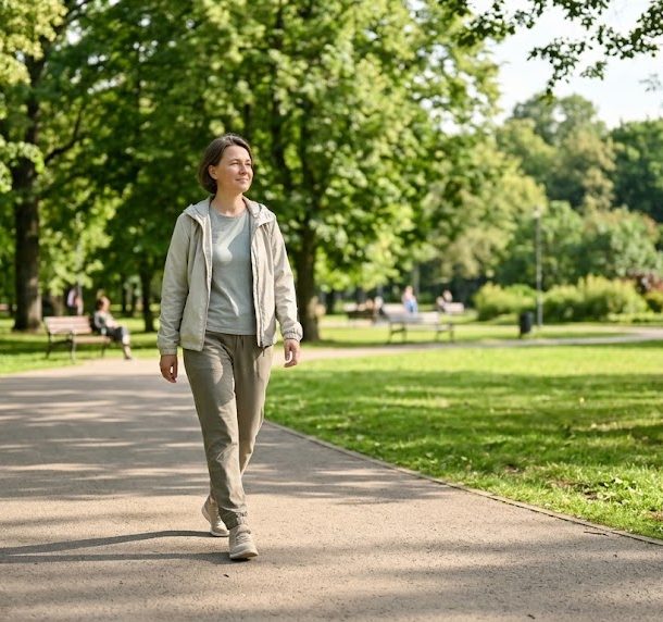 An adult in their early 40s strolling peacefully along a paved park trail on a bright, sunny afternoon. Wearing casual outdoor attire like a light jacket and comfortable pants, they have a gentle, content expression as they walk. The paved trail curves through a lush public park filled with mature green trees, open grassy areas, and dappled sunlight. In the background, other park-goers and distant foliage are softly out of focus under the warm, natural daylight. Camera: natural candid depth of field.
